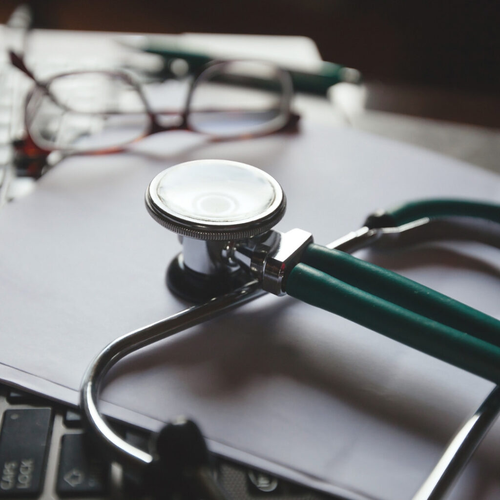 Close-up of a stethoscope resting on a stack of papers with a pair of eyeglasses in the background, suggesting a clinical setting.