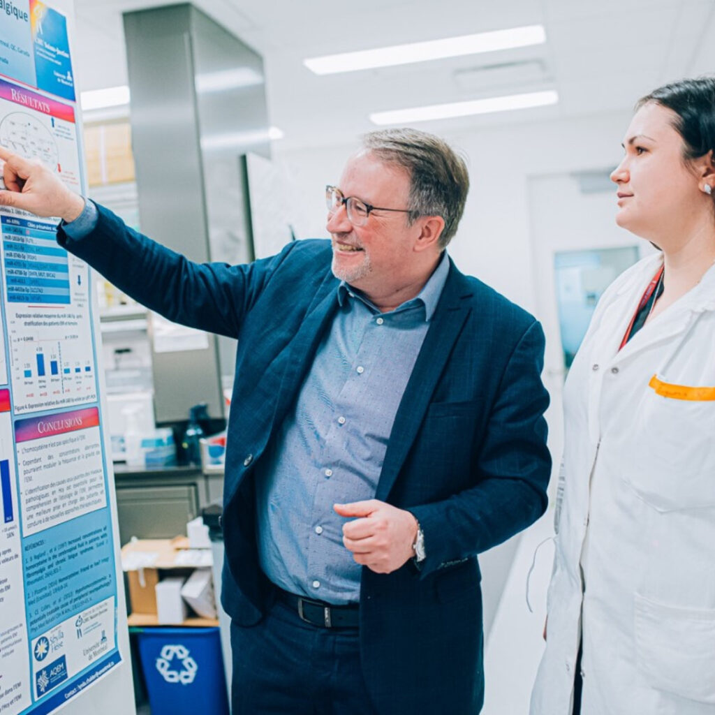 Dr. Moreau in a suit points to charts on a scientific poster while his colleague in a lab coat looks on in a laboratory.