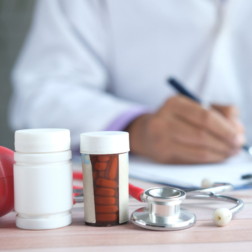 Healthcare provider writing on a chart, with a stethoscope and various pills and medicines visible in front of him.