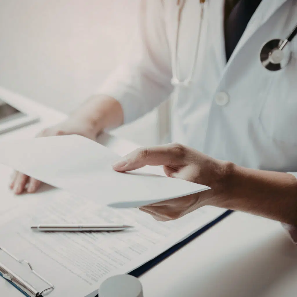 A healthcare professional in a white coat and stethoscope reviewing documents on a clipboard at a desk.